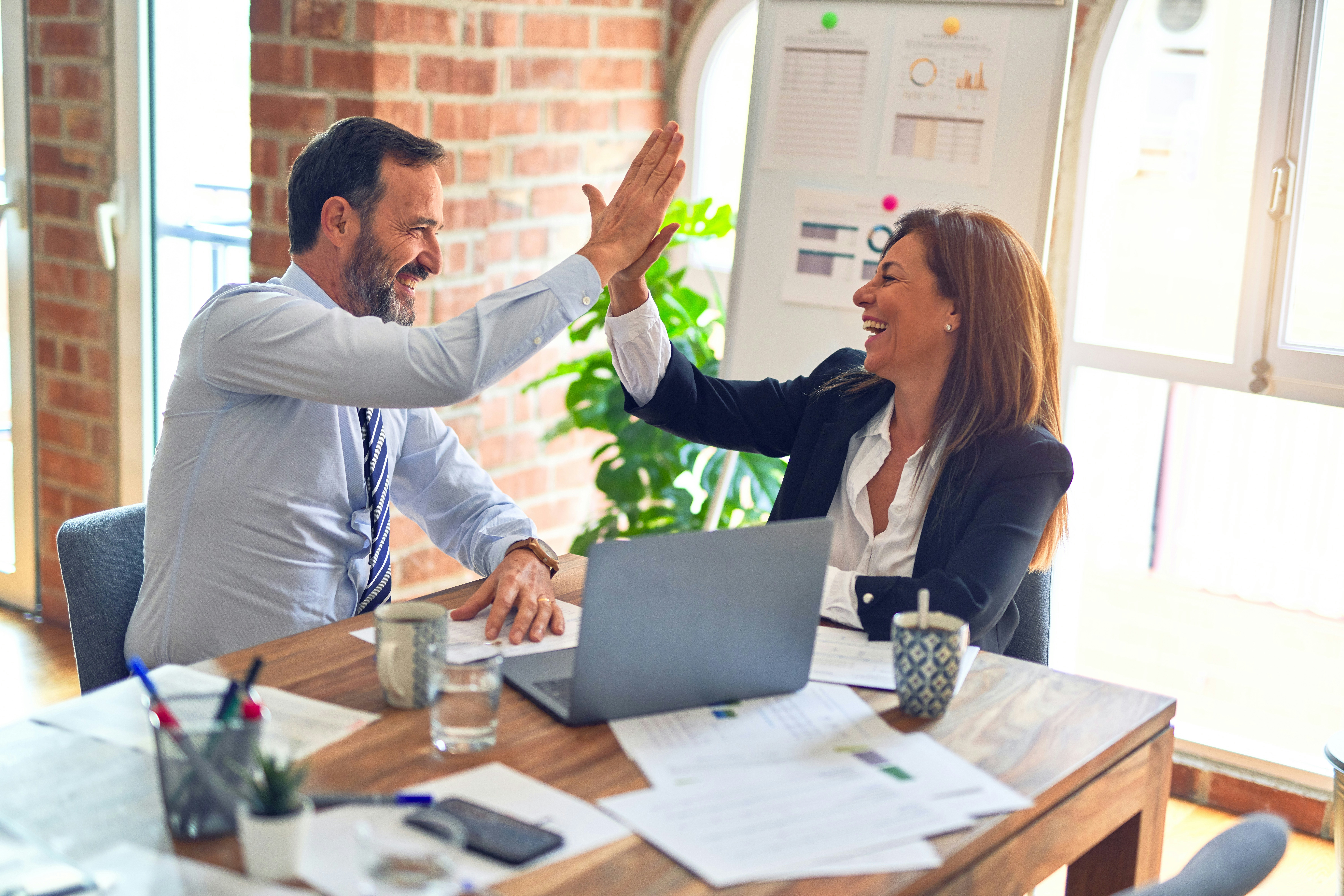 A man in white dress shirt sitting beside a woman in black long sleeve shirt working together at an office conference table and giving each other a high five